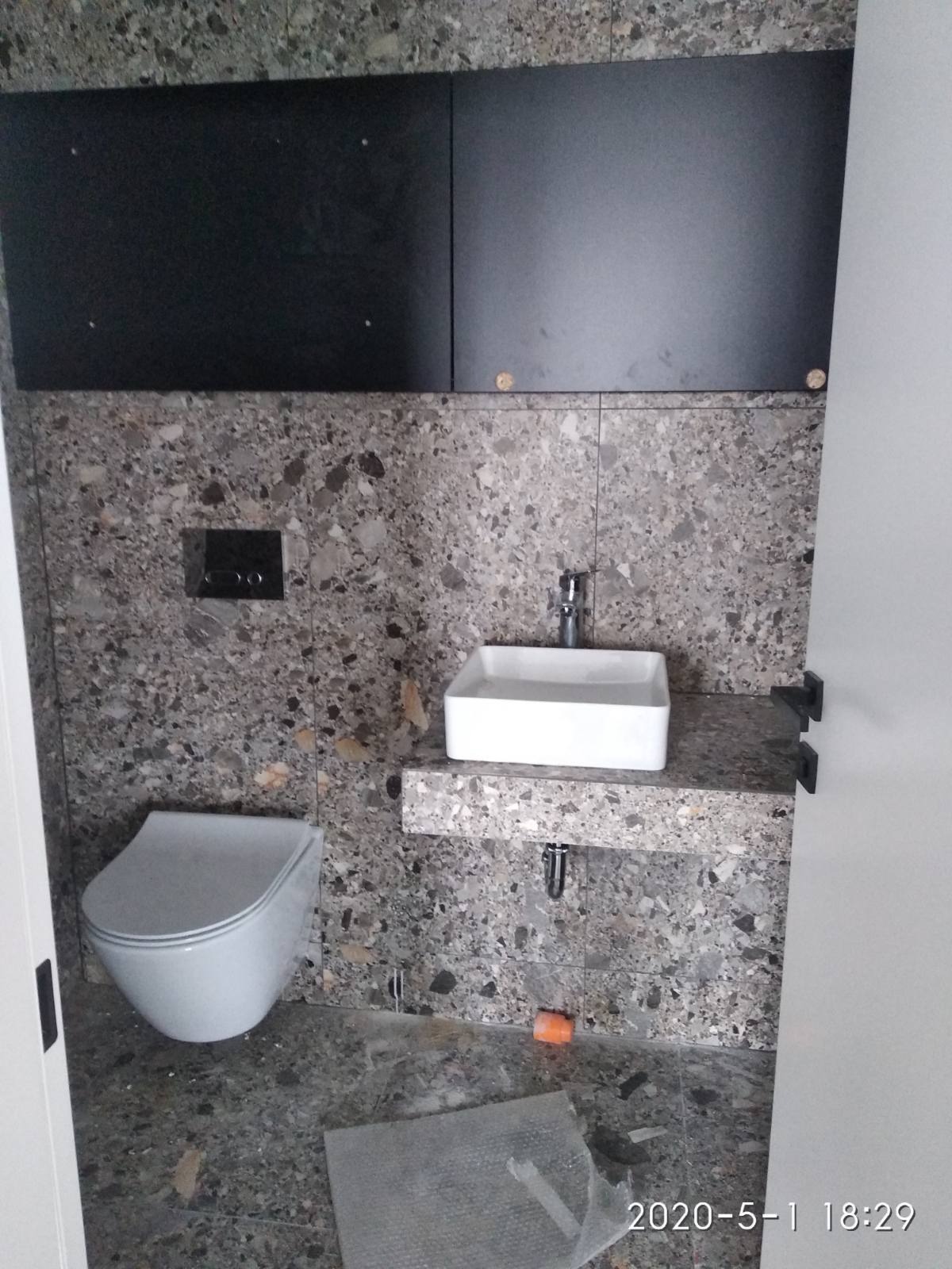 Nowoczesna łazienka z szarą mozaikową okładziną ścian i podłogi, wisząca toaleta, kwadratowa umywalka na blacie z tego samego materiału i czarne, wiszące szafki.