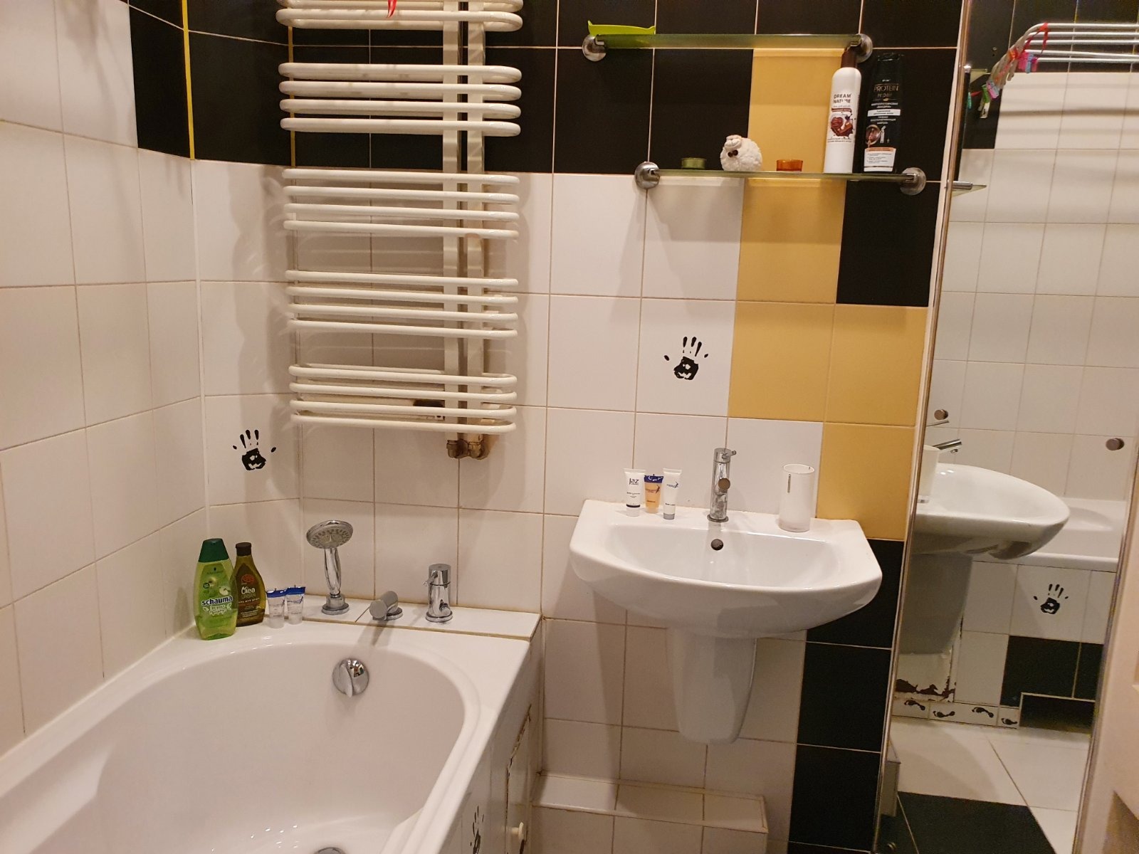 Umywalka na postumencie, wanna i grzejnik drabinkowy w łazience z biało-czarno-żółtymi płytkami i naklejkami dłoni