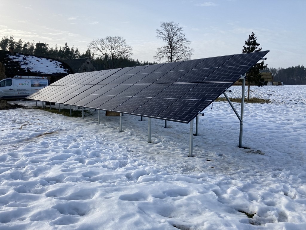 Rząd paneli słonecznych na metalowej konstrukcji, zamontowany na zasypanym śniegiem polu, z widocznym na horyzoncie budynkiem gospodarczym i samochodem dostawczym z logo firmy.