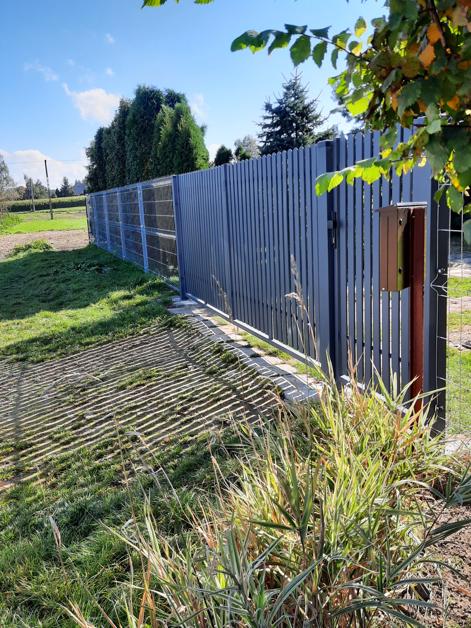 Nowoczesne ogrodzenie panelowe z metalową siatką i furtką z pionowych profili, skrzynka na listy w kolorze brązowym, cień rzucany przez ogrodzenie na trawnik, roślinność ozdobna na pierwszym planie.