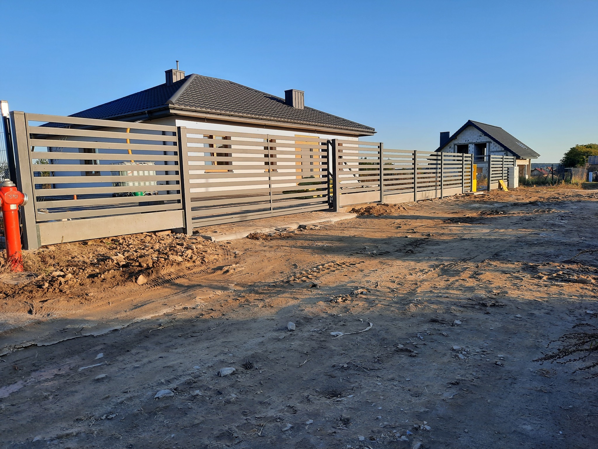 Nowoczesne, poziome ogrodzenie panelowe w kolorze szarym, oddzielające posesję z dwoma domami, jeden w stanie surowym, drugi wykończony, na tle błękitnego nieba.