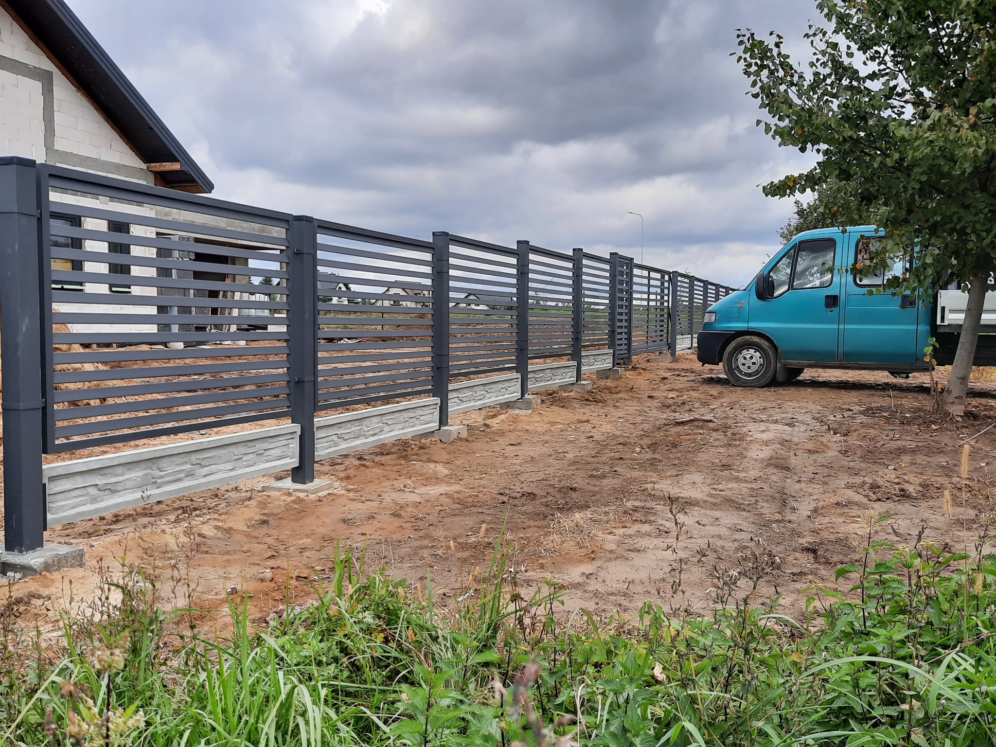 Nowoczesne, poziome ogrodzenie panelowe w kolorze grafitowym z podmurówką betonową o fakturze kamienia, widoczne na tle budowy domu i zaparkowanego niebieskiego samochodu dostawczego.