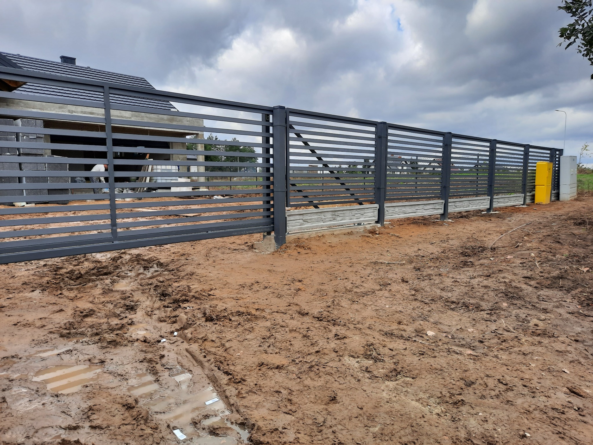 Nowoczesne, poziome przęsła w grafitowym kolorze tworzą ogrodzenie posesji, osadzone na betonowej podmurówce z dekoracyjnym wzorem, widoczne ślady po pracach ziemnych.