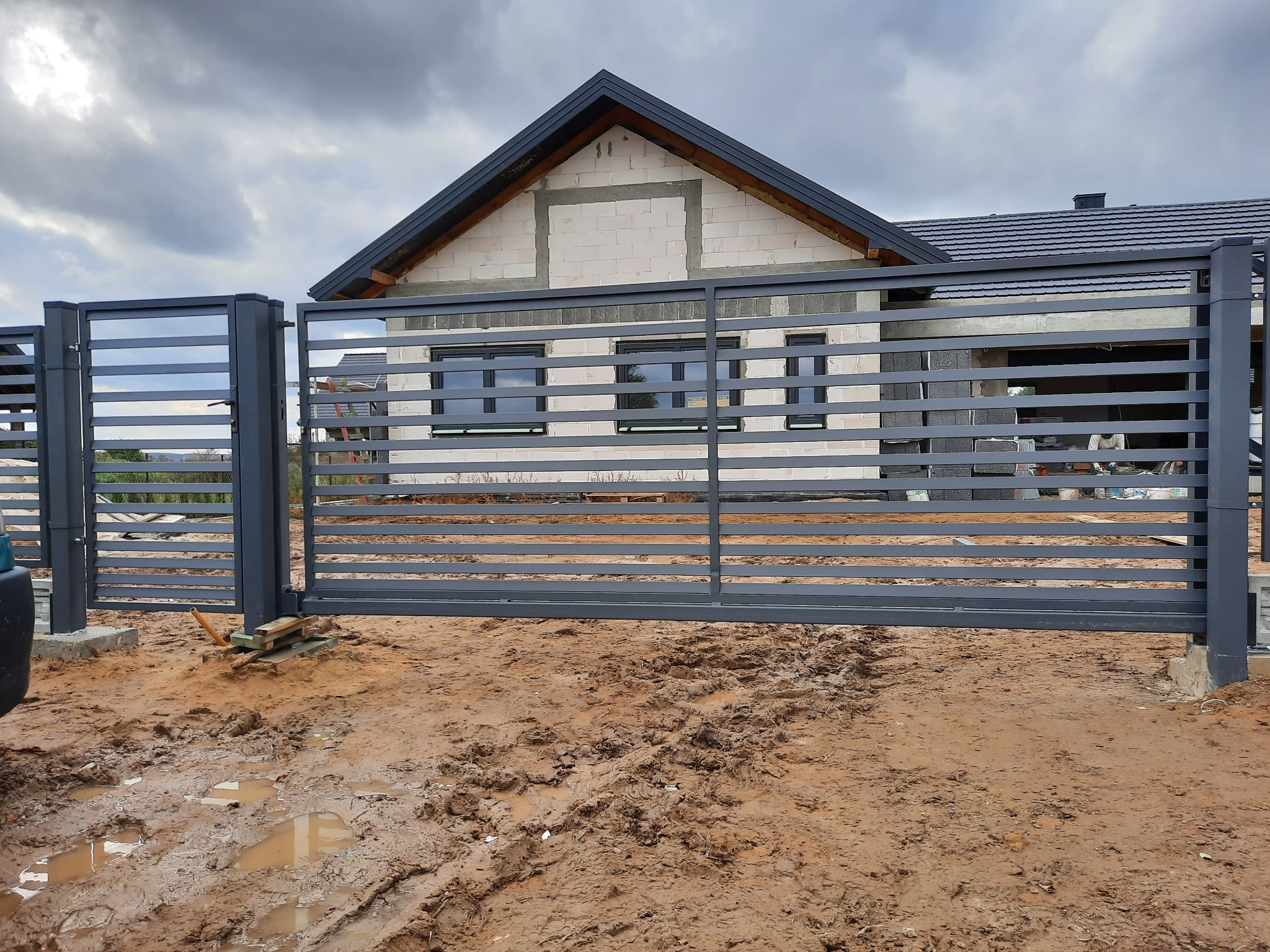 Nowoczesna, pozioma, metalowa brama przesuwna w kolorze grafitowym, zainstalowana przed domem w trakcie budowy z błotnistym terenem.