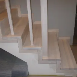 Jasne, drewniane schody z prostą balustradą w trakcie montażu, zabezpieczone folią malarską u dołu ściany i podłogi.