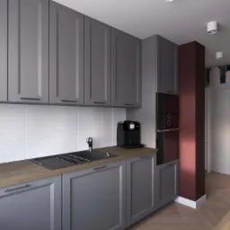 Szara kuchnia z drewnianym blatem, białymi płytkami i ciemnoczerwoną kolumną, widok na podłogę ułożoną w jodełkę i dekoracyjną tapetę w tle.