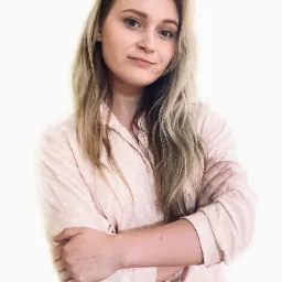 Portret młodej kobiety z blond włosami, ubranej w różową koszulę w kropki i dżinsy, z założonymi rękami, na jasnym tle.