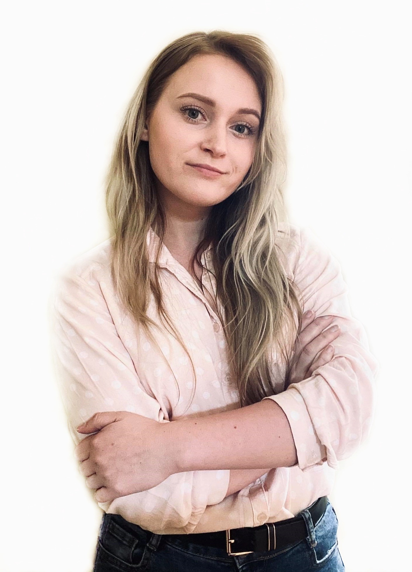 Portret młodej kobiety z blond włosami, ubranej w różową koszulę w kropki i dżinsy, z założonymi rękami, na jasnym tle.
