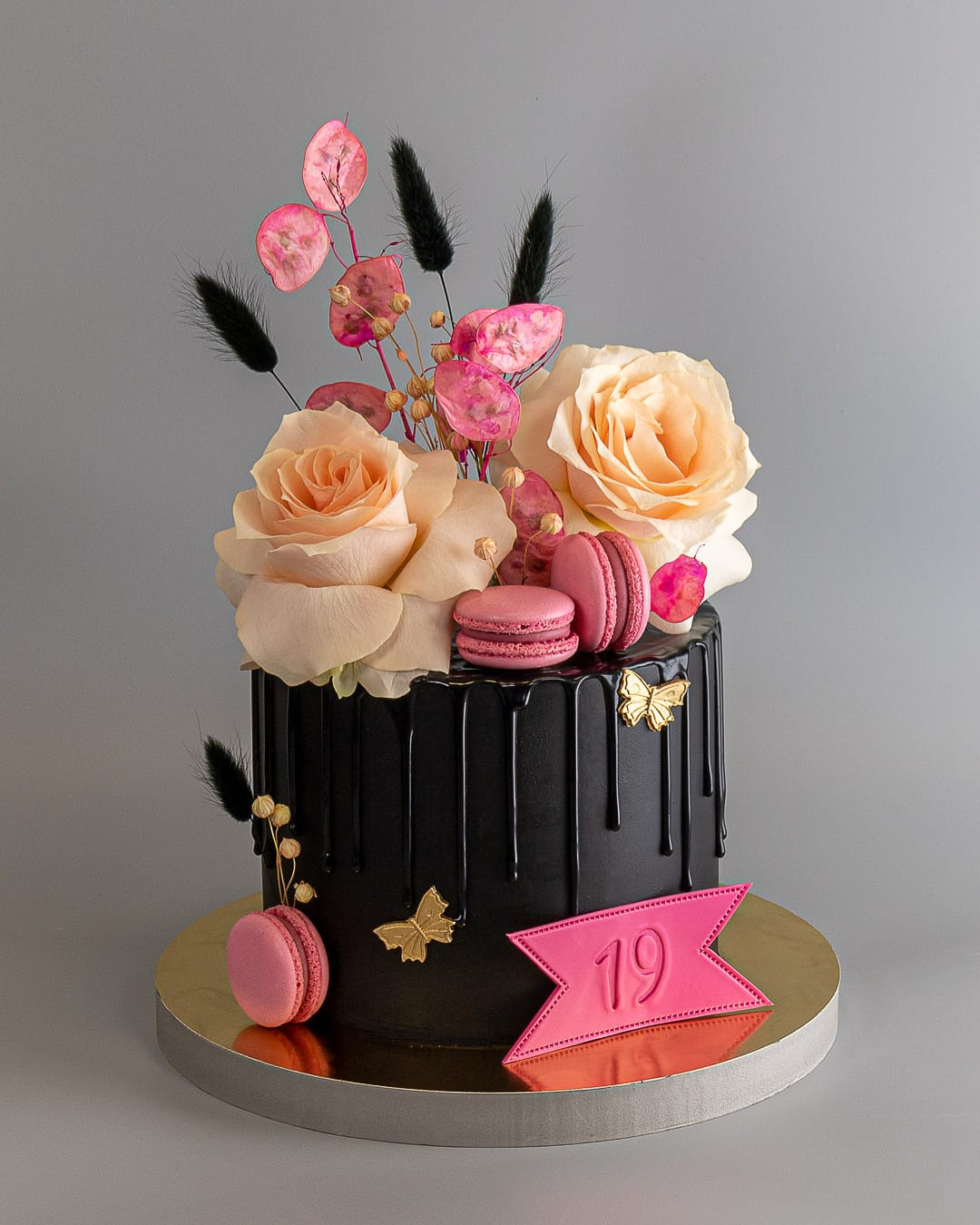 Czarny tort urodzinowy z płynną polewą, różowymi makaronikami, kwiatami i dekoracją z numerem 19 na złotej podstawie.