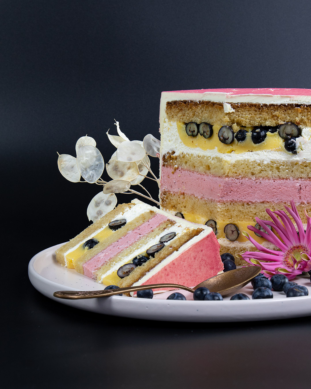 Przekrój wielowarstwowego tortu z kremem, borówkami i dekoracją z suszonych kwiatów na talerzu z ciemnym tłem.