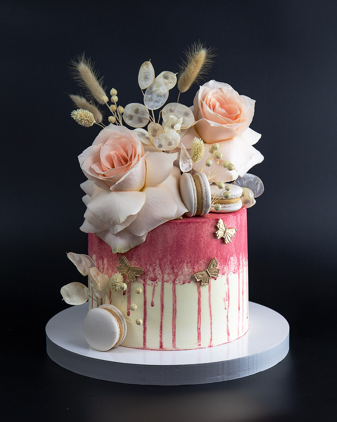 Tort z kremem ombre w odcieniach różu i bieli, udekorowany świeżymi różami, makaronikami i złotymi motylami na czarnym tle.