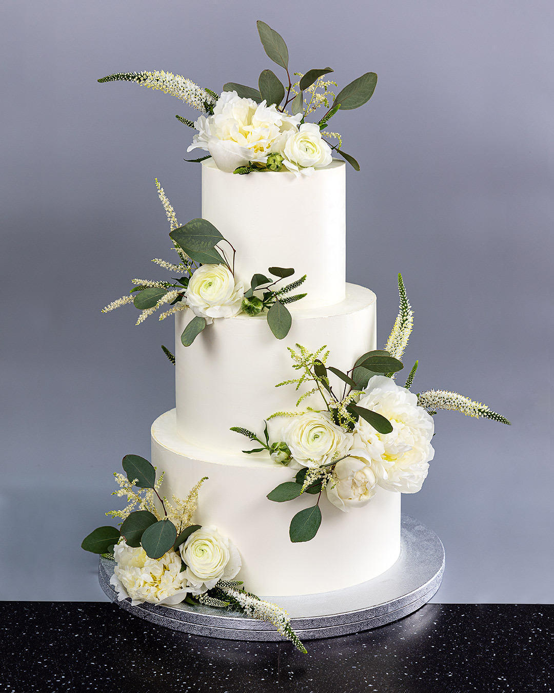 Elegancki, trzypiętrowy tort weselny w kolorze białym, udekorowany świeżymi białymi kwiatami i zielonymi liśćmi eukaliptusa na srebrnej podstawie.