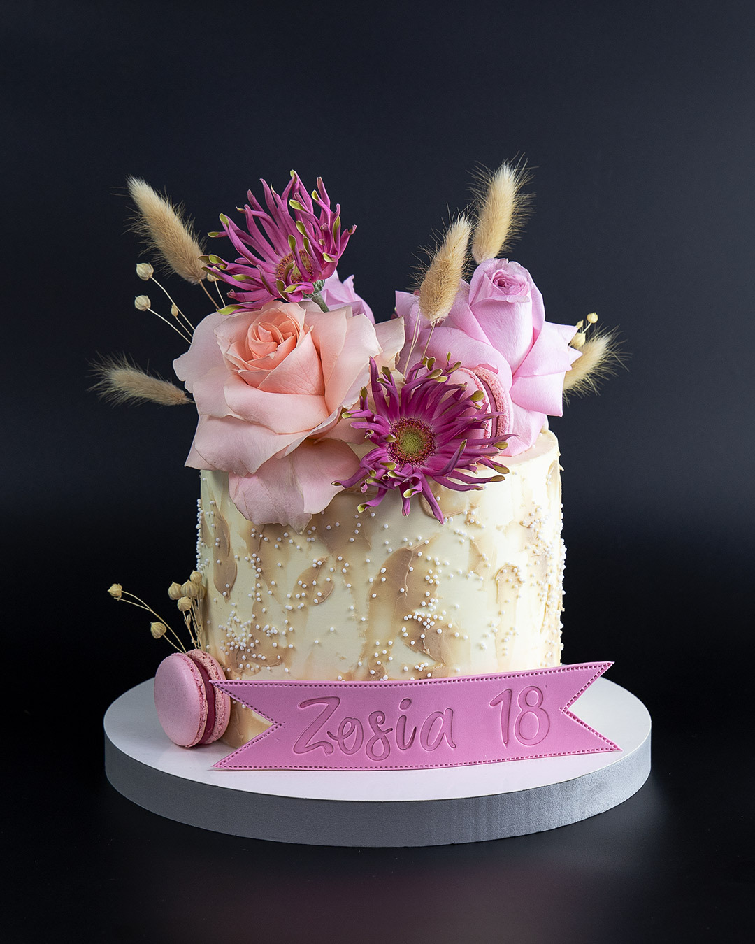 Tort zdobiony różowymi różami, fioletowymi chryzantemami i makaronikami, z różową wstążką z napisem 'Zosia 18' na białym, okrągłym podeście.