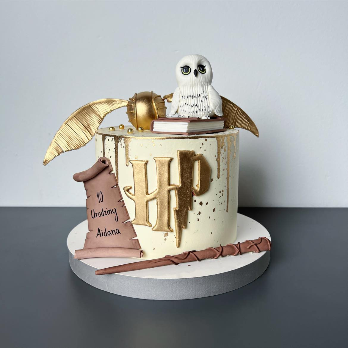 Tort urodzinowy w stylu Harry'ego Pottera z figurką sowy na książce, złotym zniczem i pergaminem z napisem '10 Urodziny Aidana', na białym tle z szarym blatem.