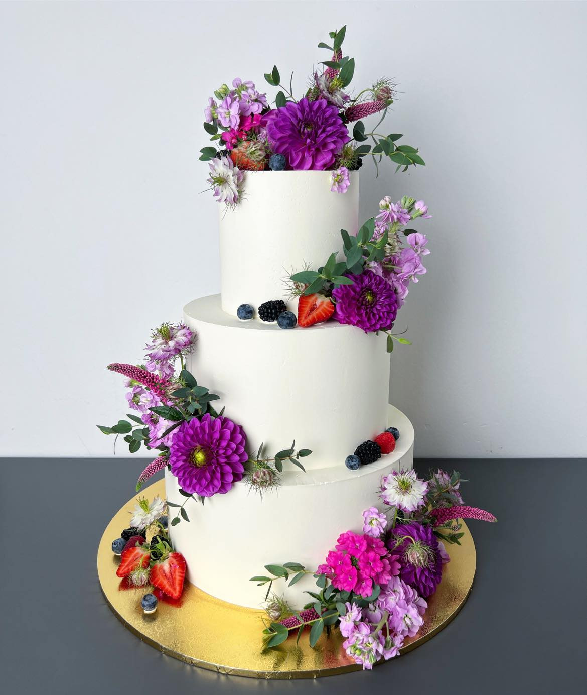 Trzypiętrowy tort weselny z białym kremem, ozdobiony świeżymi, fioletowymi i różowymi kwiatami oraz owocami leśnymi na złotej podstawie.