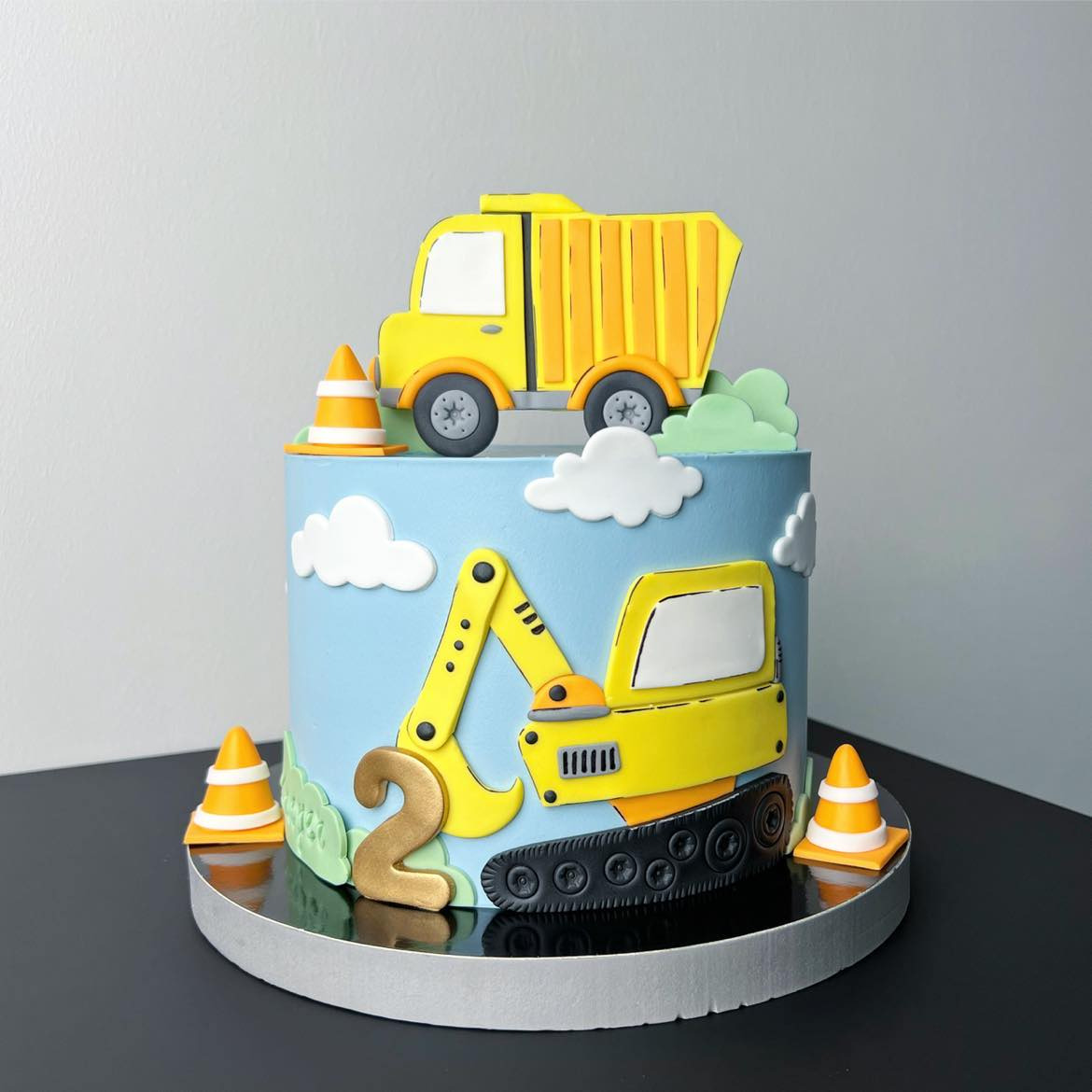 Tort urodzinowy w Poznaniu z motywem budowlanym: żółta ciężarówka na górze, koparka z boku, cyfra '2' i stożki drogowe, całość na jasnoniebieskim tle z białymi chmurkami.