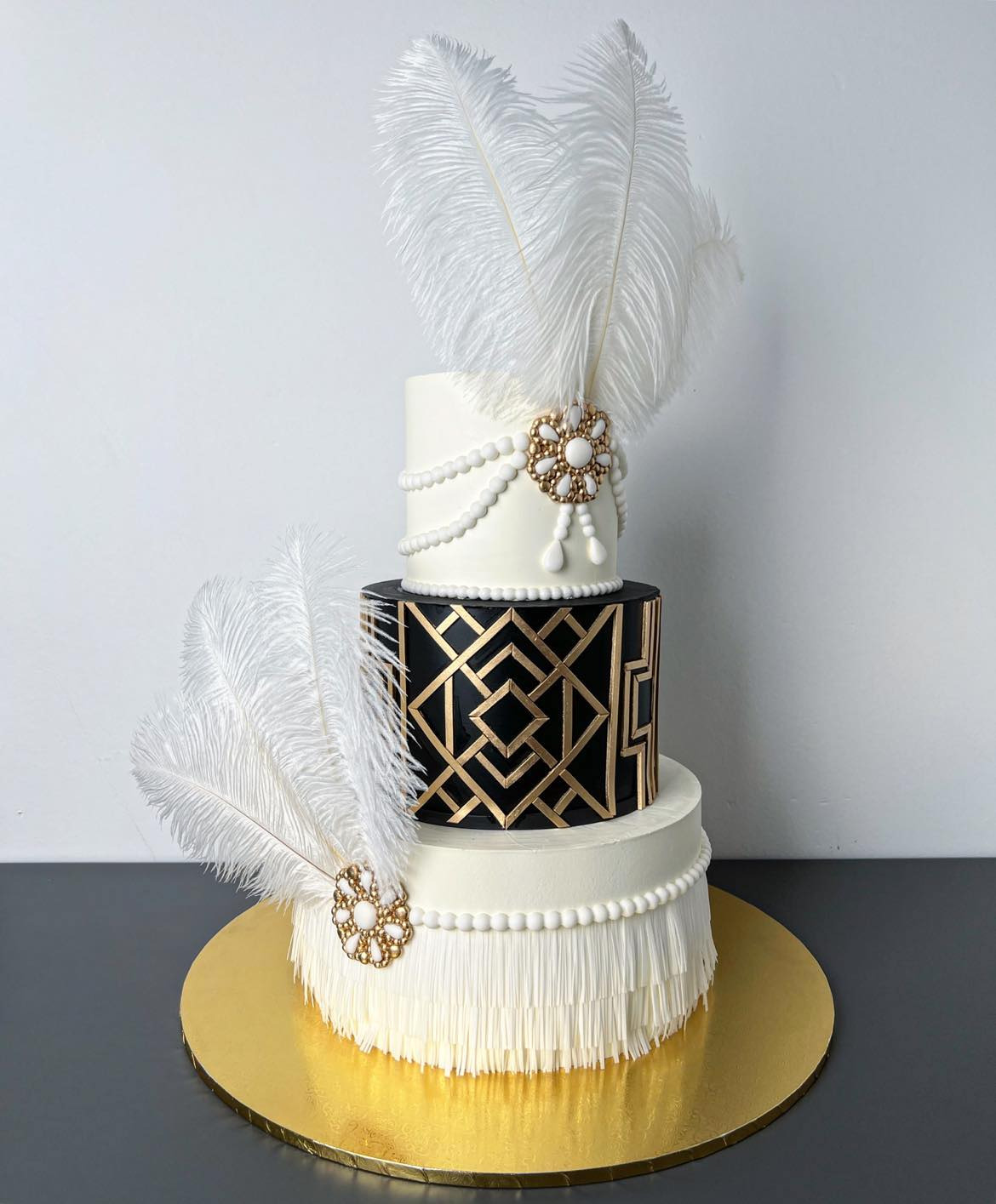 Trzypiętrowy tort w stylu Art Deco z białymi piórami, złotymi zdobieniami i perłowymi detalami na złotym podkładzie.