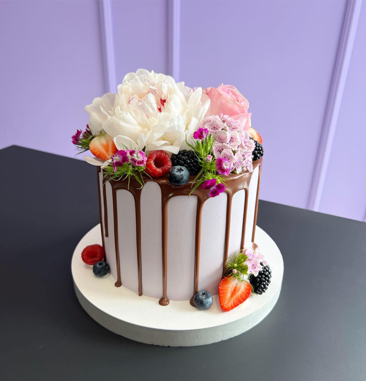Elegancki tort z białym kremem, polewą czekoladową i dekoracją z białej piwonii, różowej róży, drobnych fioletowych kwiatów, truskawek, malin i jagód, na białym podkładzie i ciemnoszarym tle.