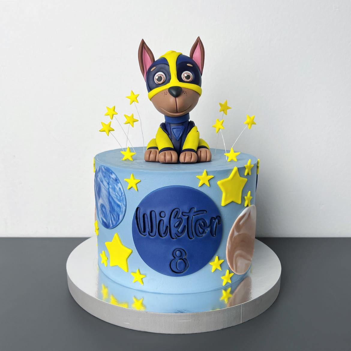 Tort urodzinowy w jasnoniebieskim kolorze z motywem kosmosu i figurką psa z bajki na wierzchu, ozdobiony żółtymi gwiazdkami i napisem 'Wiktor 8'.