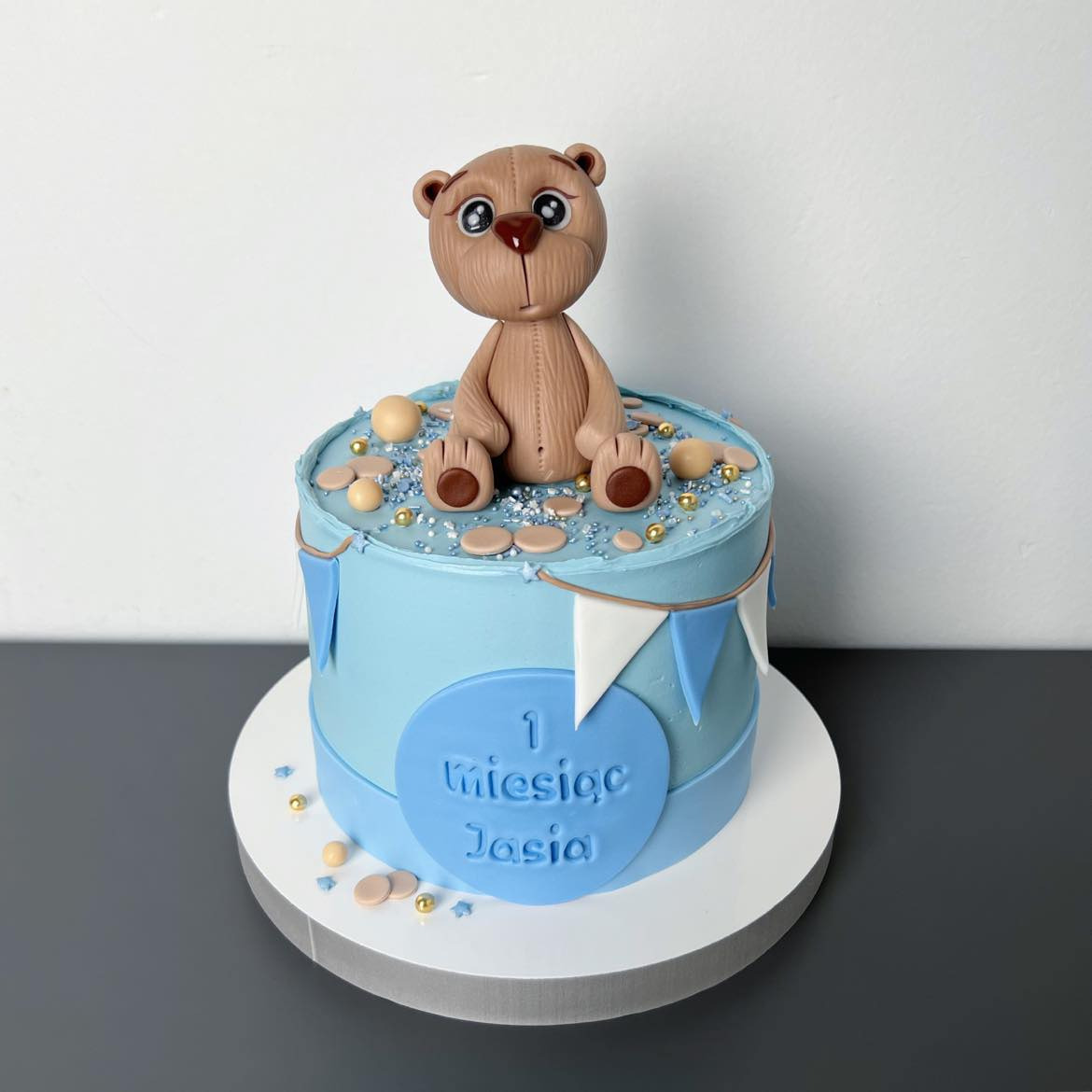 Tort na pierwsze urodziny z dekoracją w postaci misia z lukru, pastelowo-niebieski krem i napis '1 Miesiąc Jasia'.