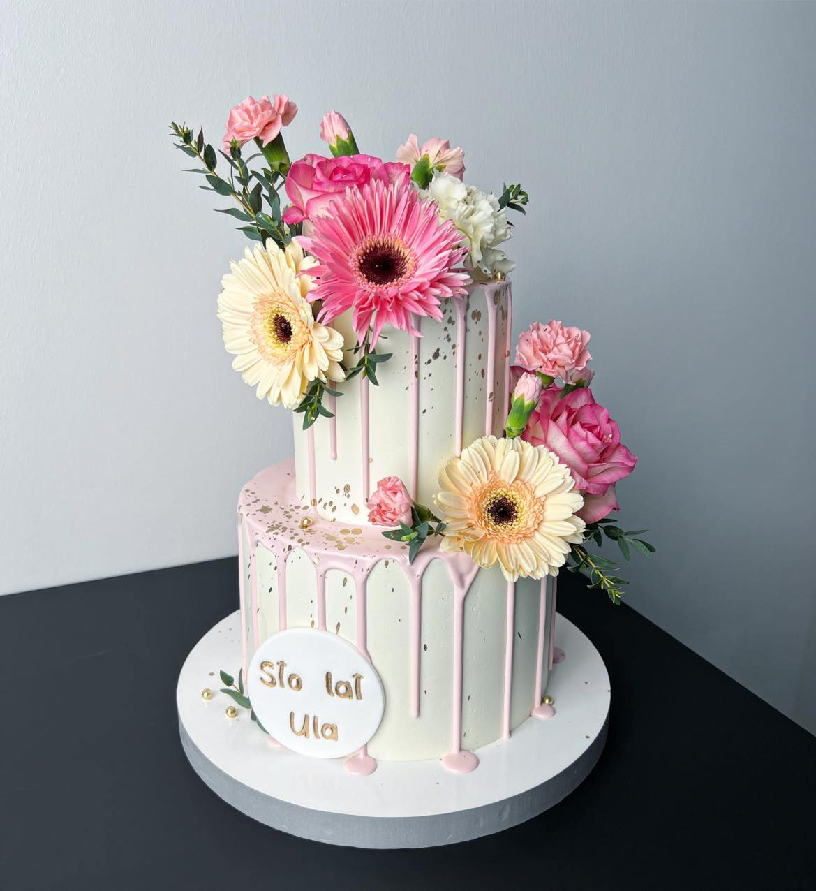 Dwupoziomowy tort urodzinowy z białym lukrem, różowym drippingiem, złotymi akcentami i dekoracją z żywych kwiatów: gerbery, róże, goździki; na podstawie okrągły napis 'Sto lat Ula'.