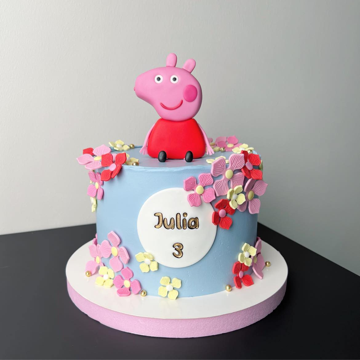 Tort urodzinowy dla Julii z figurką świnki Peppy, udekorowany pastelowymi kwiatami i złotymi kuleczkami na niebieskim tle.
