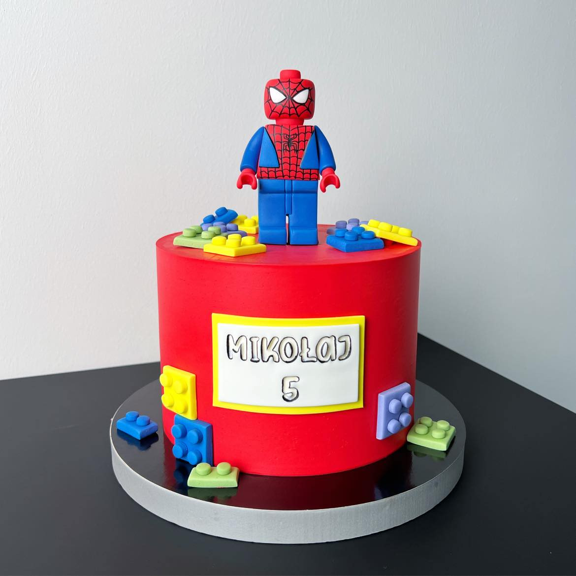 Czerwony tort urodzinowy z figurką Spider-Mana z klocków Lego i napisem 'Mikołaj 5', otoczony kolorowymi klockami na czarnym tle.