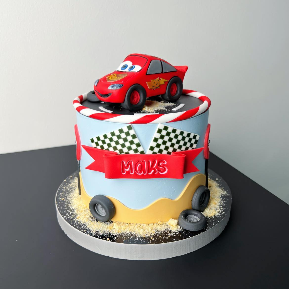 Tort urodzinowy dla dziecka z motywem wyścigowym, dekorowany figurką czerwonego auta i flagami w szachownicę, z napisem 'Maks' na czerwonej wstążce.