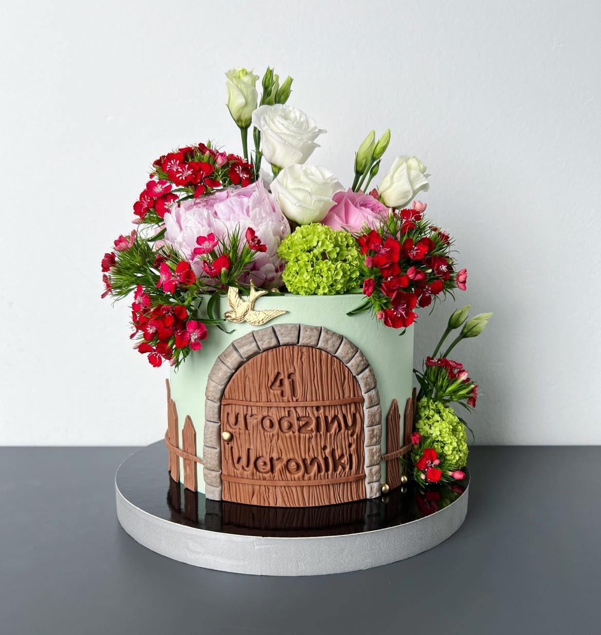 Tort urodzinowy w kształcie drzwi zdobiony świeżymi kwiatami i złotym ptaszkiem, z napisem '41 Urodziny Weroniki'.