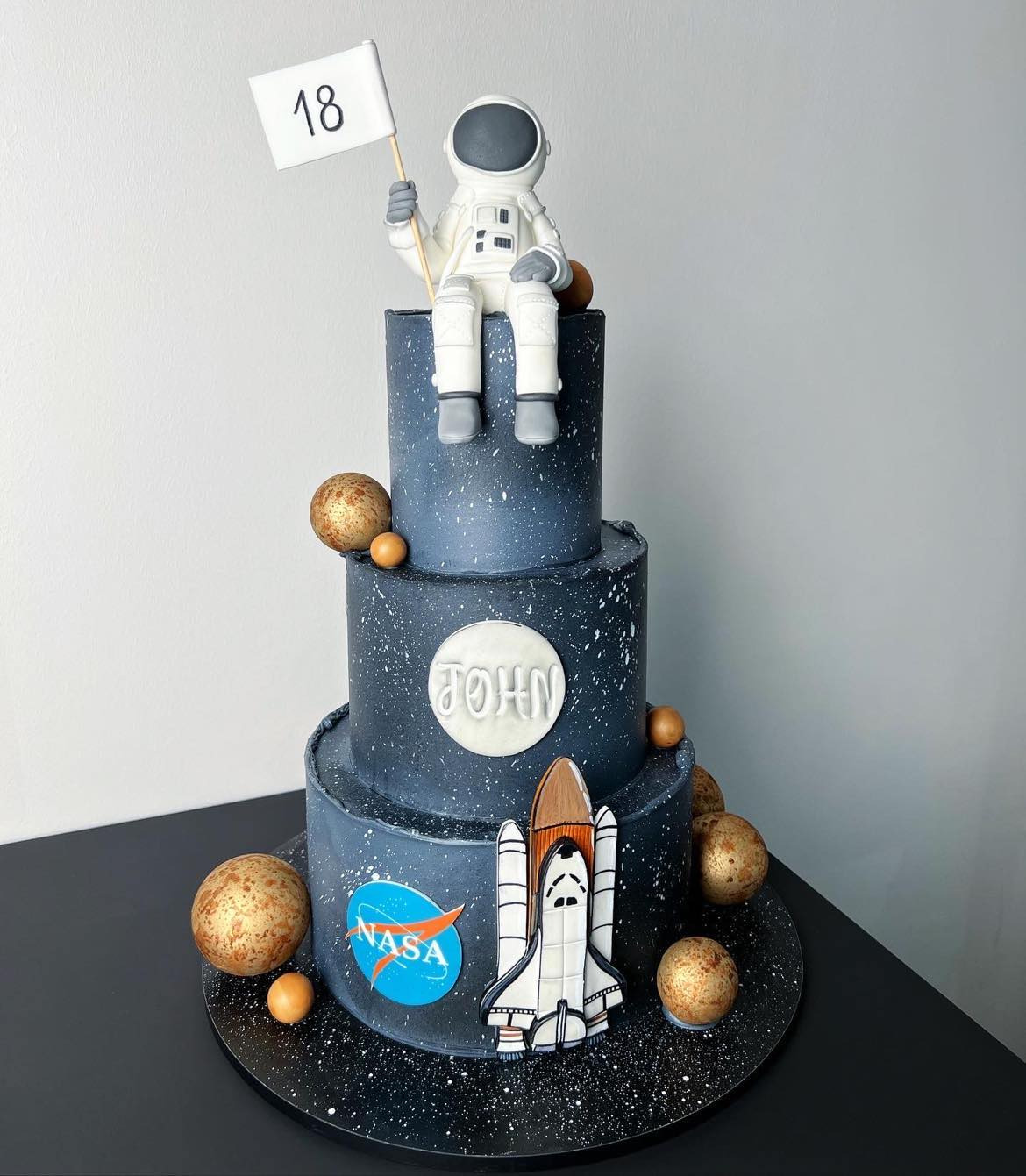 Trzypiętrowy tort urodzinowy z motywem kosmicznym: figurka astronauty z flagą '18', rakieta kosmiczna, logo NASA, planety i imię 'John' na tle ciemnogranatowej galaktyki.