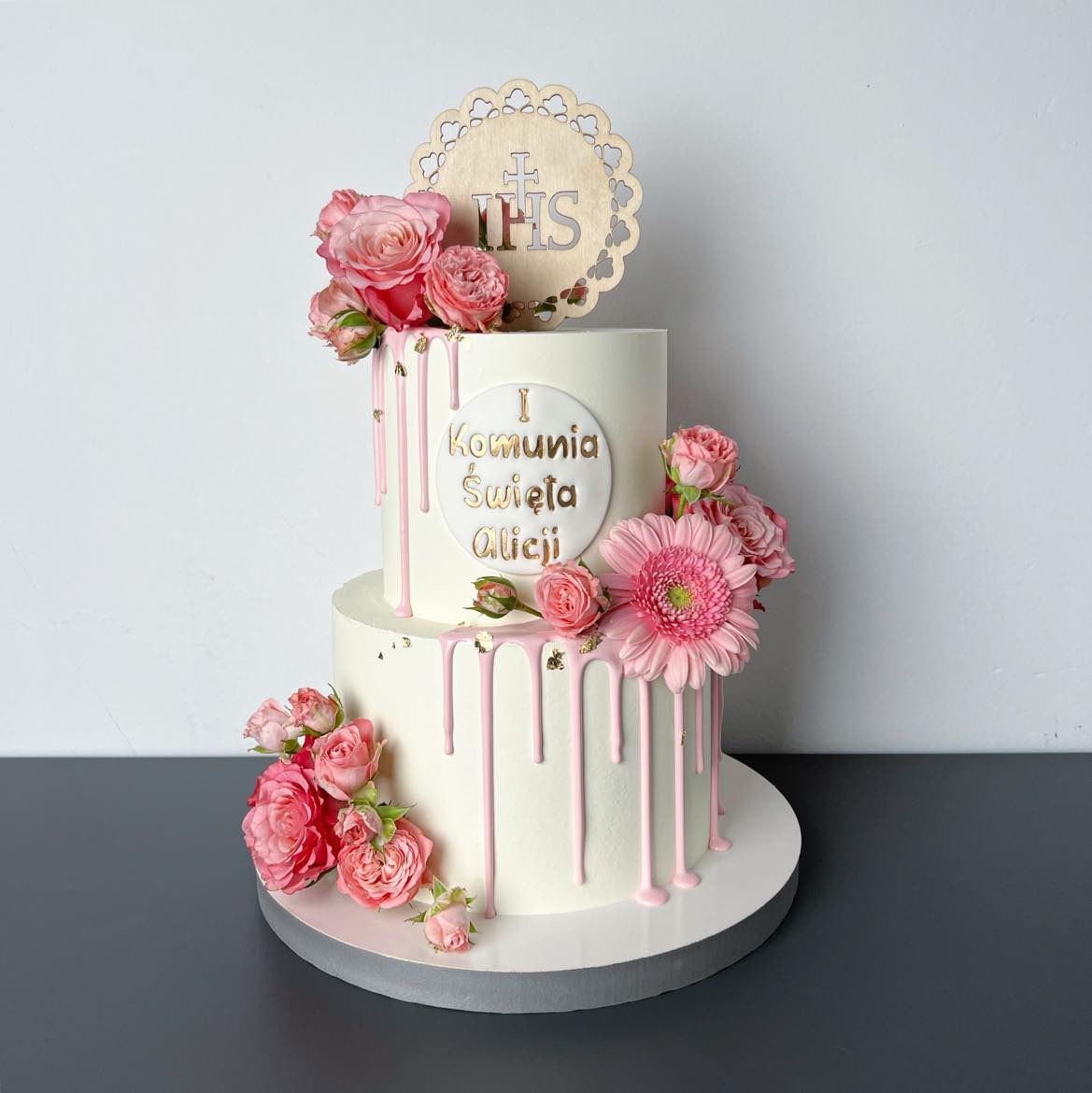 Dwupoziomowy tort komunijny z białym kremem, różowym lukrem spływającym po bokach, ozdobiony różowymi różami i gerberą, z drewnianym topperem IHS i napisem 'I Komunia Święta Alicji' w złotym kolorze.
