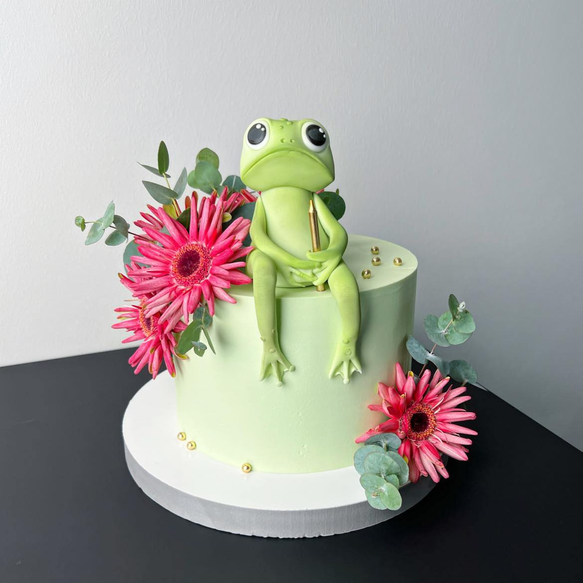 Tort z zielonym lukrem, ozdobiony figurką żaby z lukru trzymającej złotą świeczkę, otoczony różowymi gerberami i eukaliptusem.