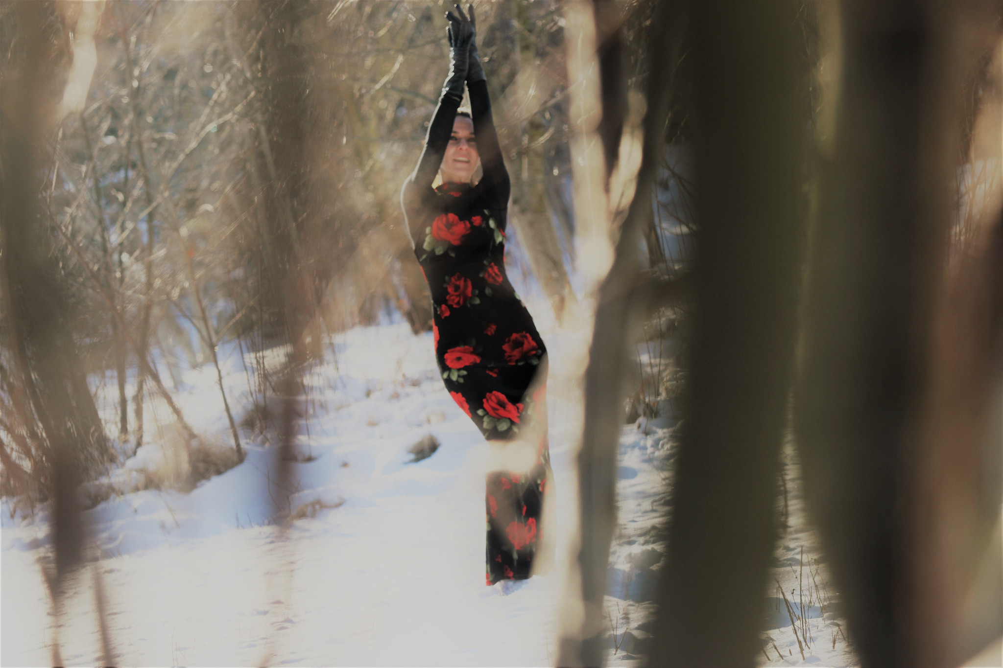 Kobieta w czarnej sukience w czerwone róże i długich rękawiczkach pozuje na śniegu w lesie, widziana przez rozmyte gałęzie na pierwszym planie.
