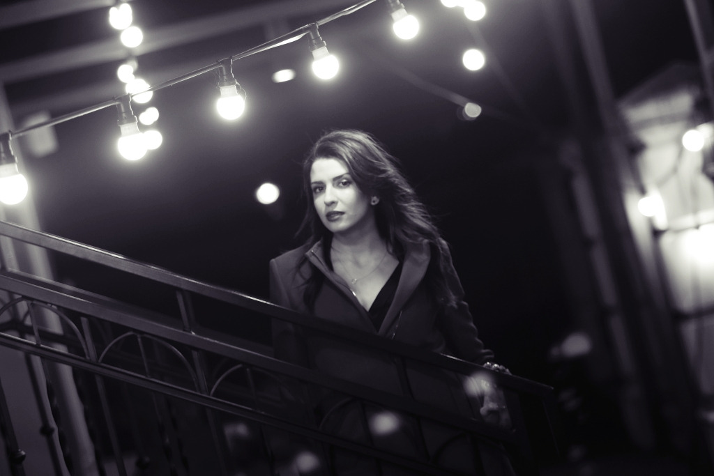 Czarno-biała fotografia kobiety w płaszczu, stojącej na schodach z metalową balustradą, oświetlonej sznurem żarówek, skierowanej w stronę obiektywu.