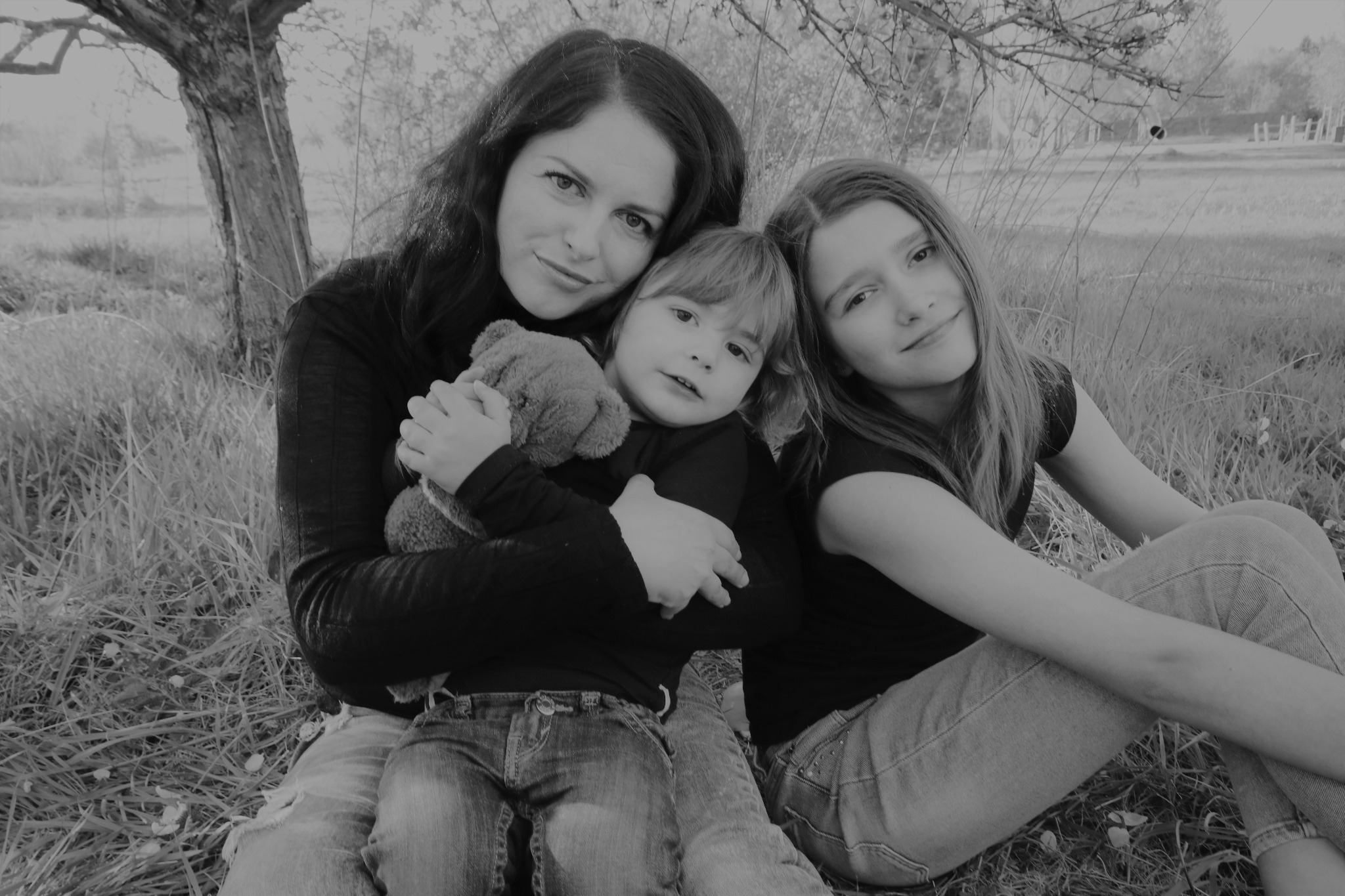 Czarno-białe zdjęcie przedstawiające matkę obejmującą dwie córki, siedzące na trawie w plenerze; młodsza dziewczynka trzyma pluszowego misia.