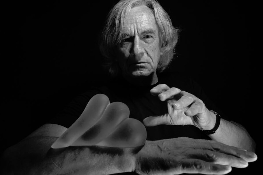 Czarno-białe portretowe ujęcie starszego mężczyzny z silikonowymi implantami piersi na dłoni, oświetlonymi z boku, na ciemnym tle.