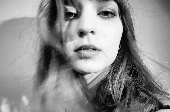 Portret kobiety w czerni i bieli, z włosami opadającymi na twarz, oświetlonej z góry, z widocznym cieniem na ścianie w tle.