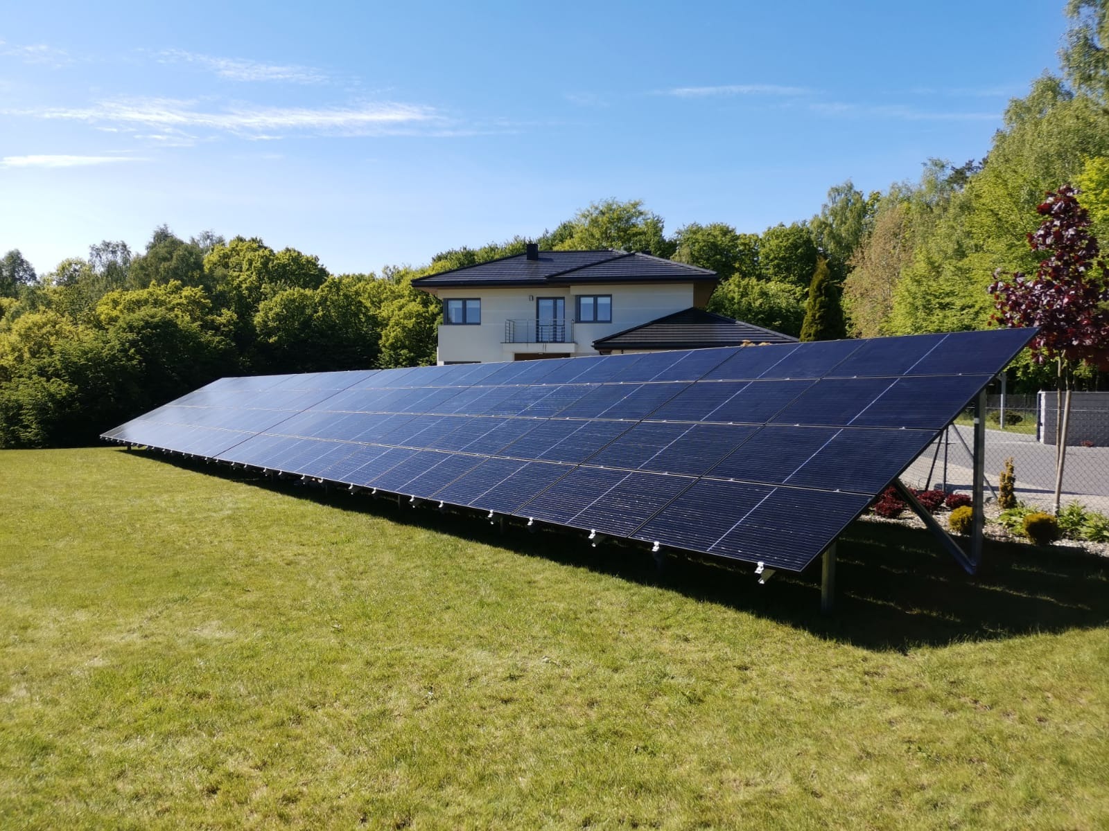 Długi szereg paneli słonecznych na trawniku przed domem jednorodzinnym, widok z niskiej perspektywy podkreślający rozległość instalacji.