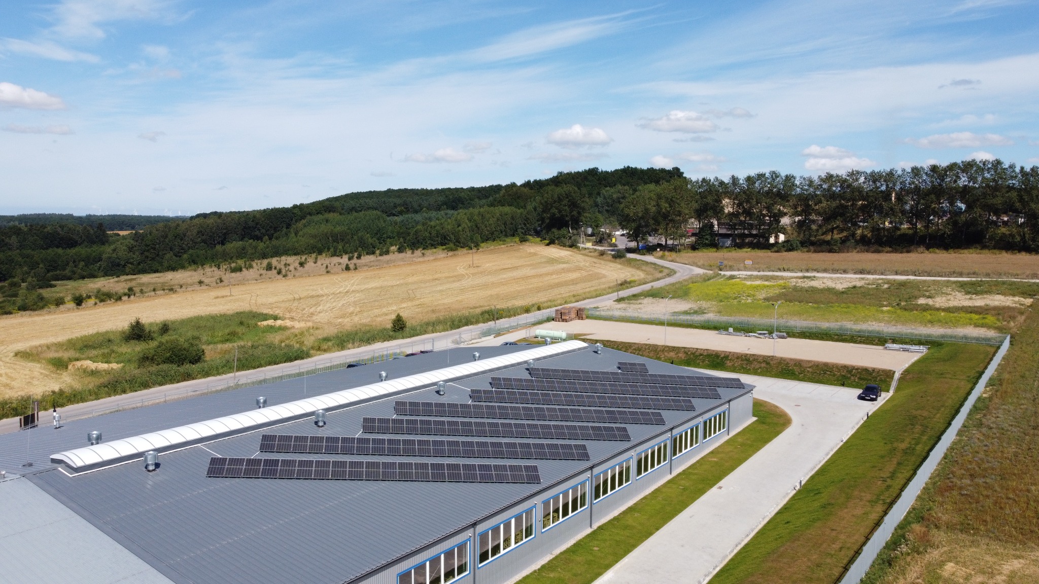 Widok z lotu ptaka na budynek przemysłowy z szarym dachem, na którym zamontowane są panele słoneczne; w tle pola, las i niebieskie niebo z chmurami.