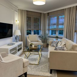 2 RAZY K - Elegancki salon z beżowymi sofami, szklanym stolikiem kawowym ze złotymi nogami i drewnianymi żaluzjami. Lampy ścienne i dekoracje dopełniają aranżację.
