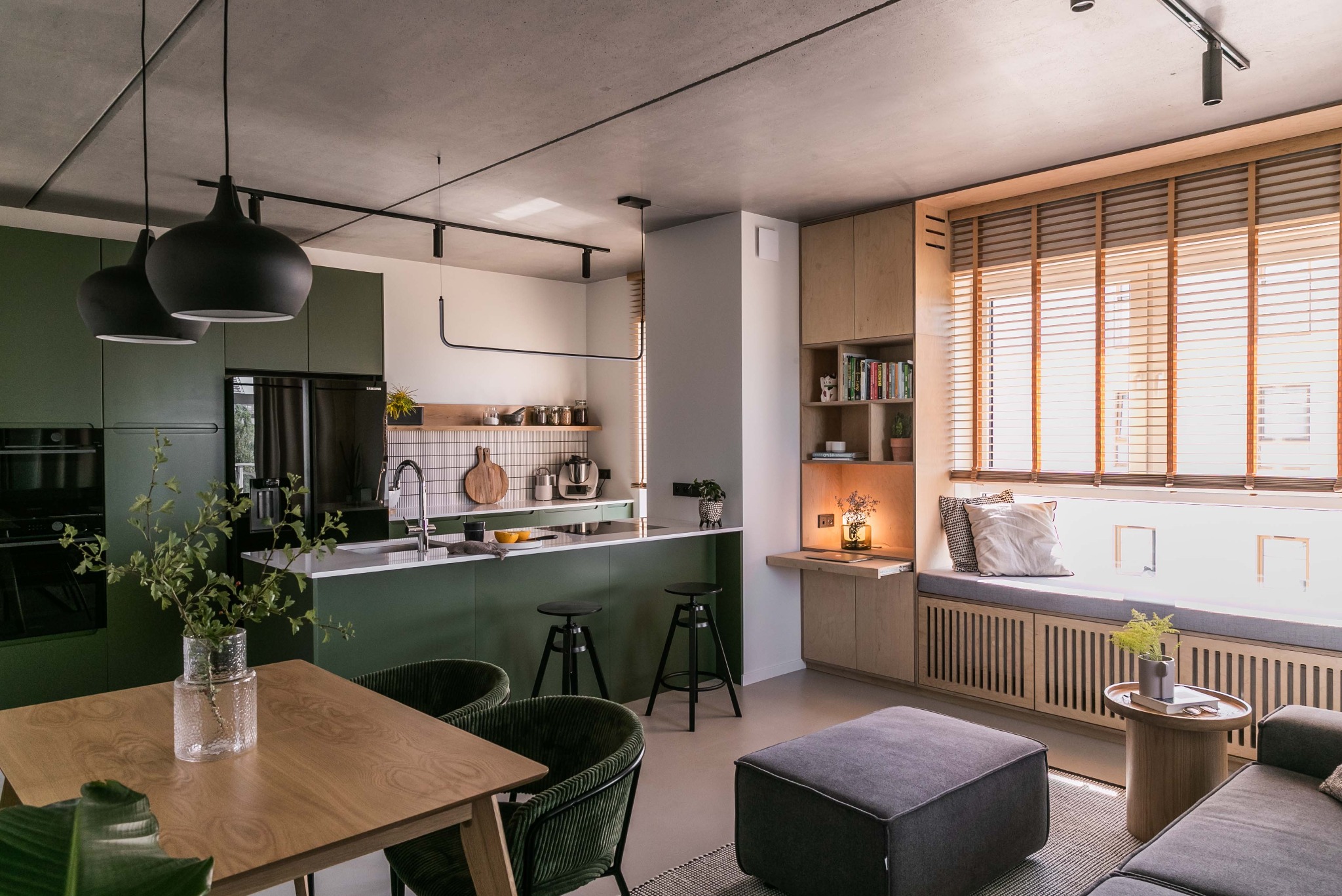 Nowoczesna kuchnia z zielonymi szafkami i wyspą, drewniany stół z zielonymi krzesłami, okno z drewnianymi żaluzjami i siedziskiem, betonowy sufit z oświetleniem szynowym.