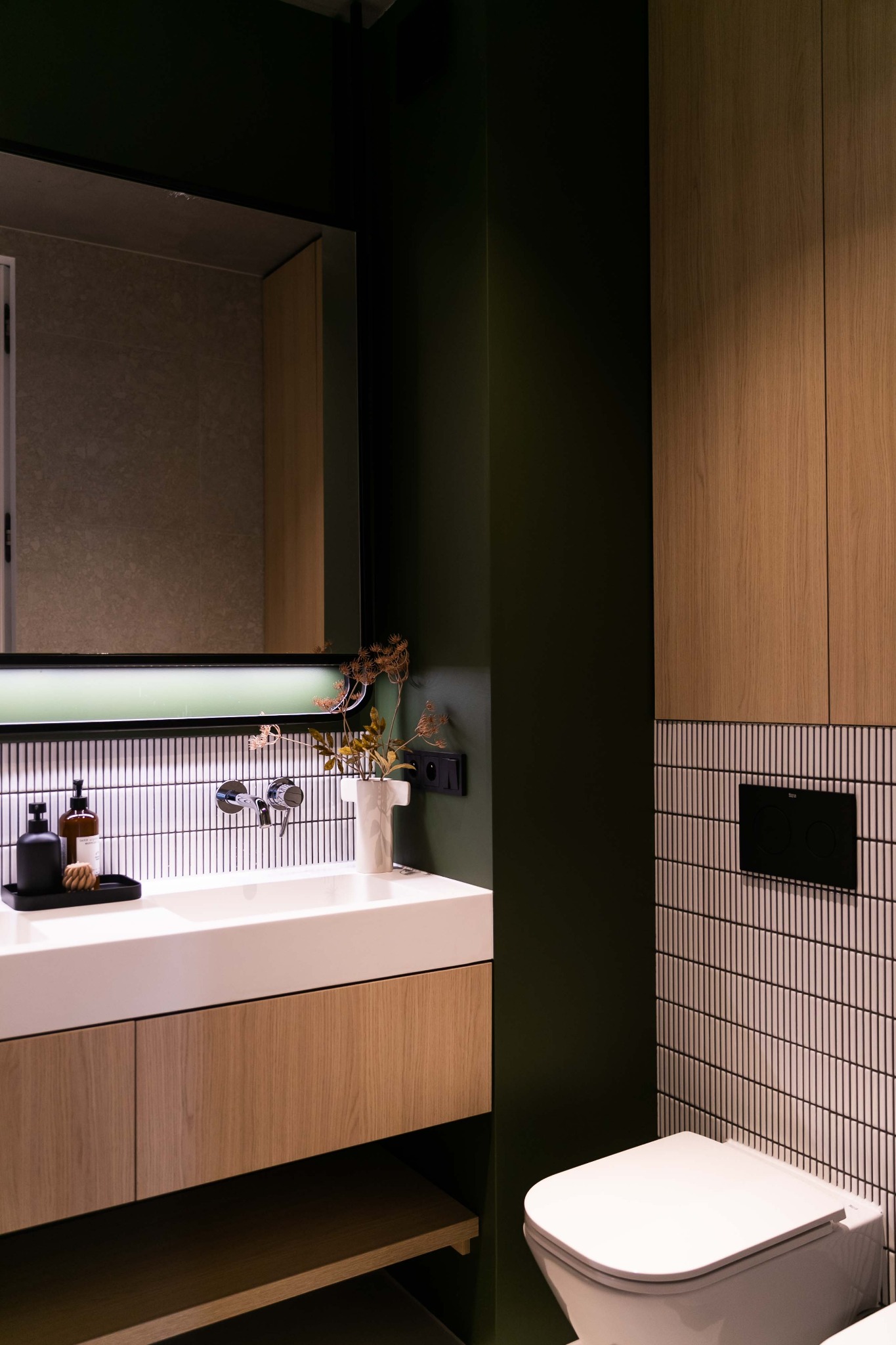 Nowoczesna łazienka z umywalką nablatową, drewnianą szafką pod umywalką i toaletą. Ściany pomalowane na ciemną zieleń i wyłożone białymi, pionowymi płytkami. Na umywalce czarne akcesoria łazienkowe...