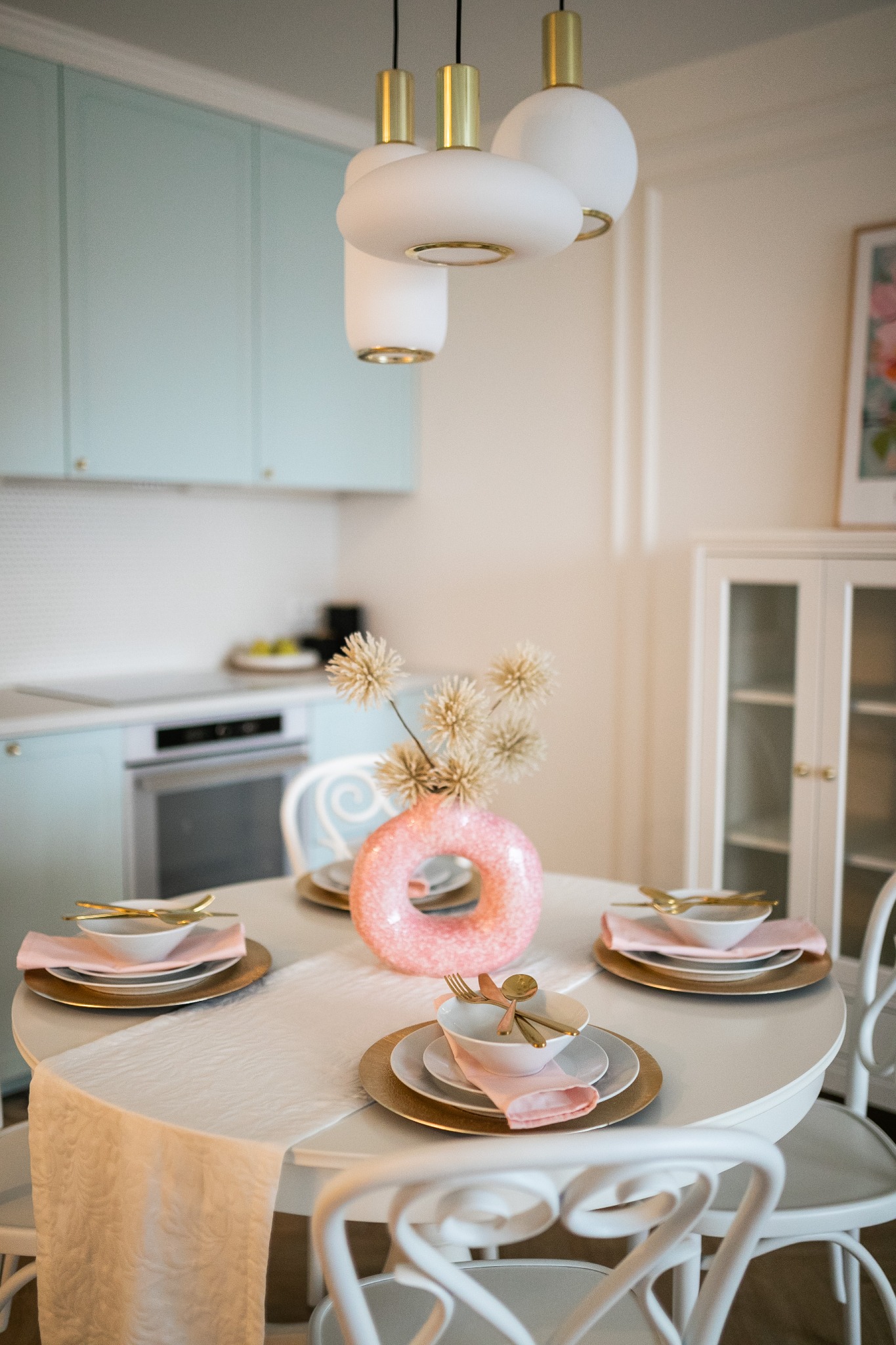 Pastelowa kuchnia z okrągłym stołem nakrytym złotą zastawą, różowymi serwetkami i oryginalnym wazonem w kształcie donuta, oświetlona nowoczesną lampą wiszącą.