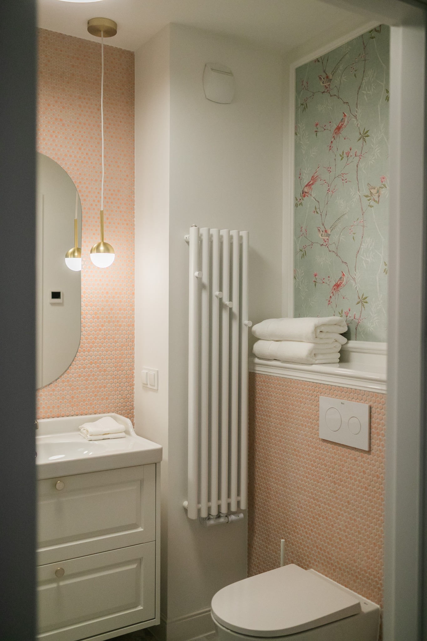 Jasna łazienka z umywalką na szafce, grzejnikiem drabinkowym, toaletą i ścianą w pastelowe mozaiki oraz fragmentem ściany z tapetą w motywy roślinne i ptasie.