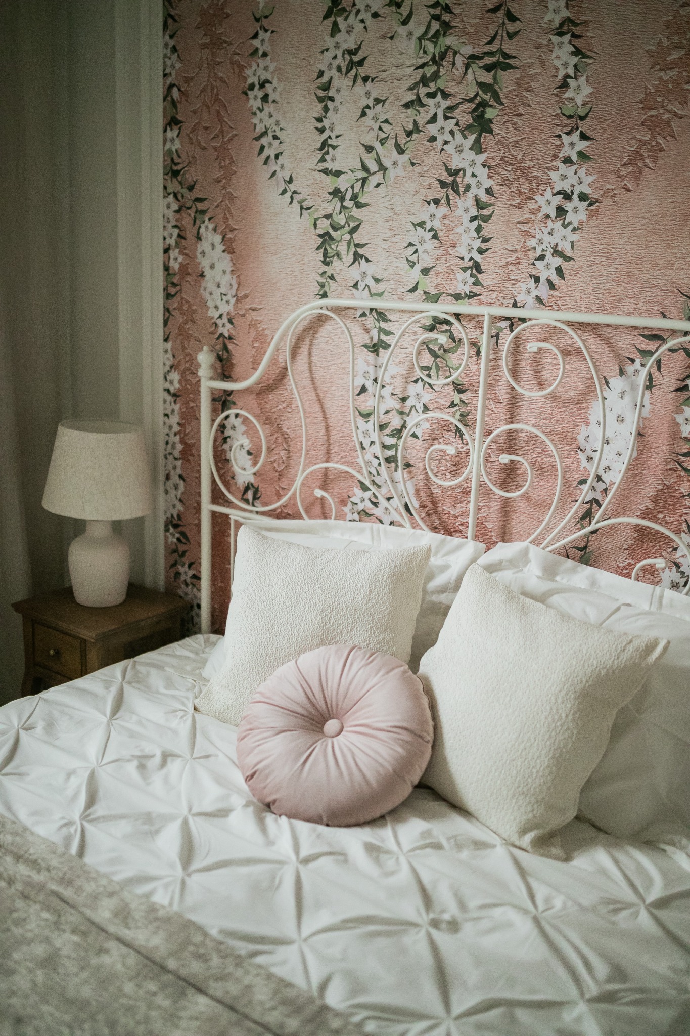 Ustawienie łóżka z kutym, białym zagłówkiem na tle ściany z tapetą w kwiaty, obejmujące białą pościel z pikowaniem, dwie białe poduszki i jedną okrągłą, pudrowo różową poduszkę.