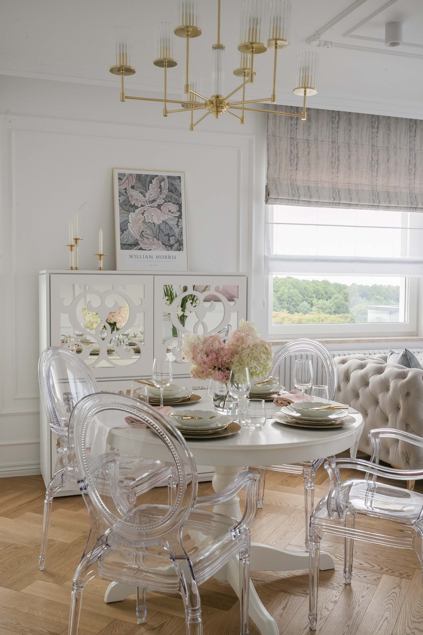 Elegancka jadalnia w jasnych barwach: okrągły stół z nakryciem, przezroczyste krzesła, komoda z lustrzanymi wstawkami i plakatem William Morrisa, złoty żyrandol.