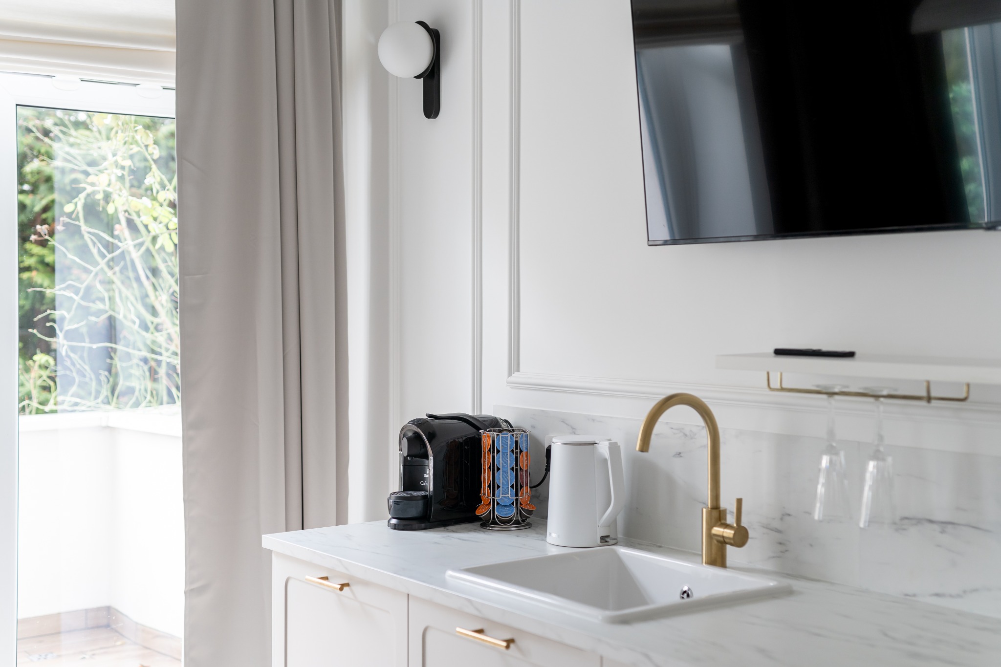 Nowoczesna aranżacja aneksu kuchennego z marmurowym blatem, złotą armaturą i ekspres do kawy, widok na balkon z zielenią w tle.