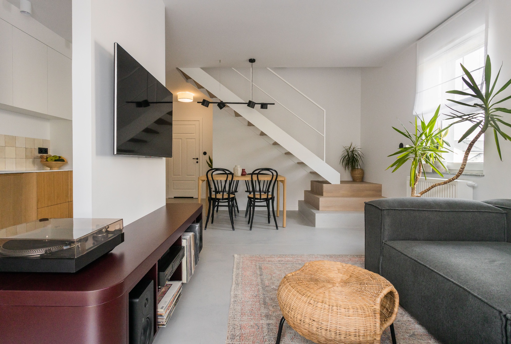 Jasne, minimalistyczne wnętrze z szarą sofą, drewnianym stolikiem RTV z gramofonem, schodami z białymi barierkami, drewnianym stołem z czarnymi krzesłami i roślinami doniczkowymi.