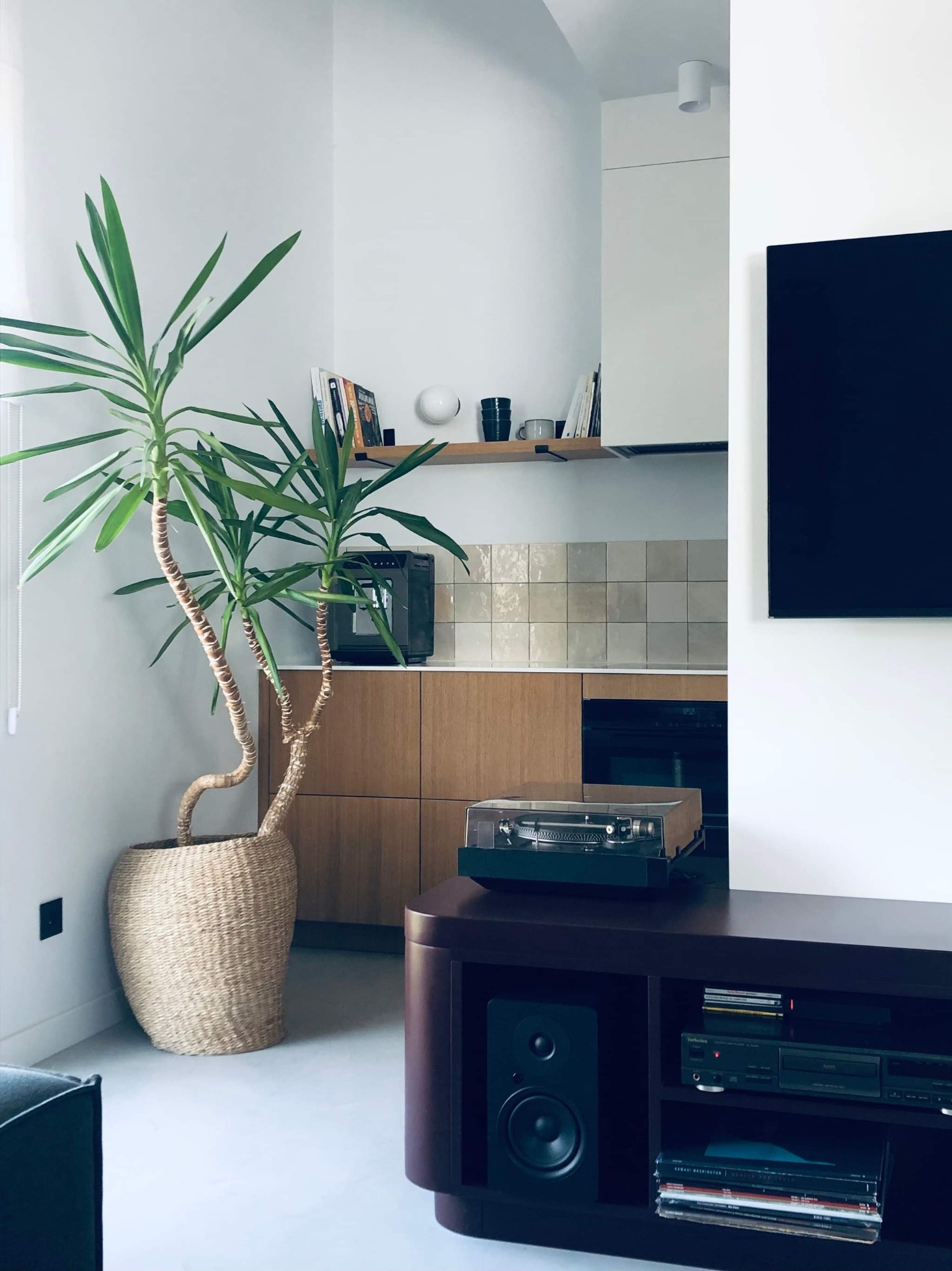 Jasne wnętrze mieszkania z rośliną w wiklinowym koszu, gramofonem na ciemnej komodzie, oraz widocznym fragmentem kuchni z drewnianymi szafkami i płytkami na ścianie.