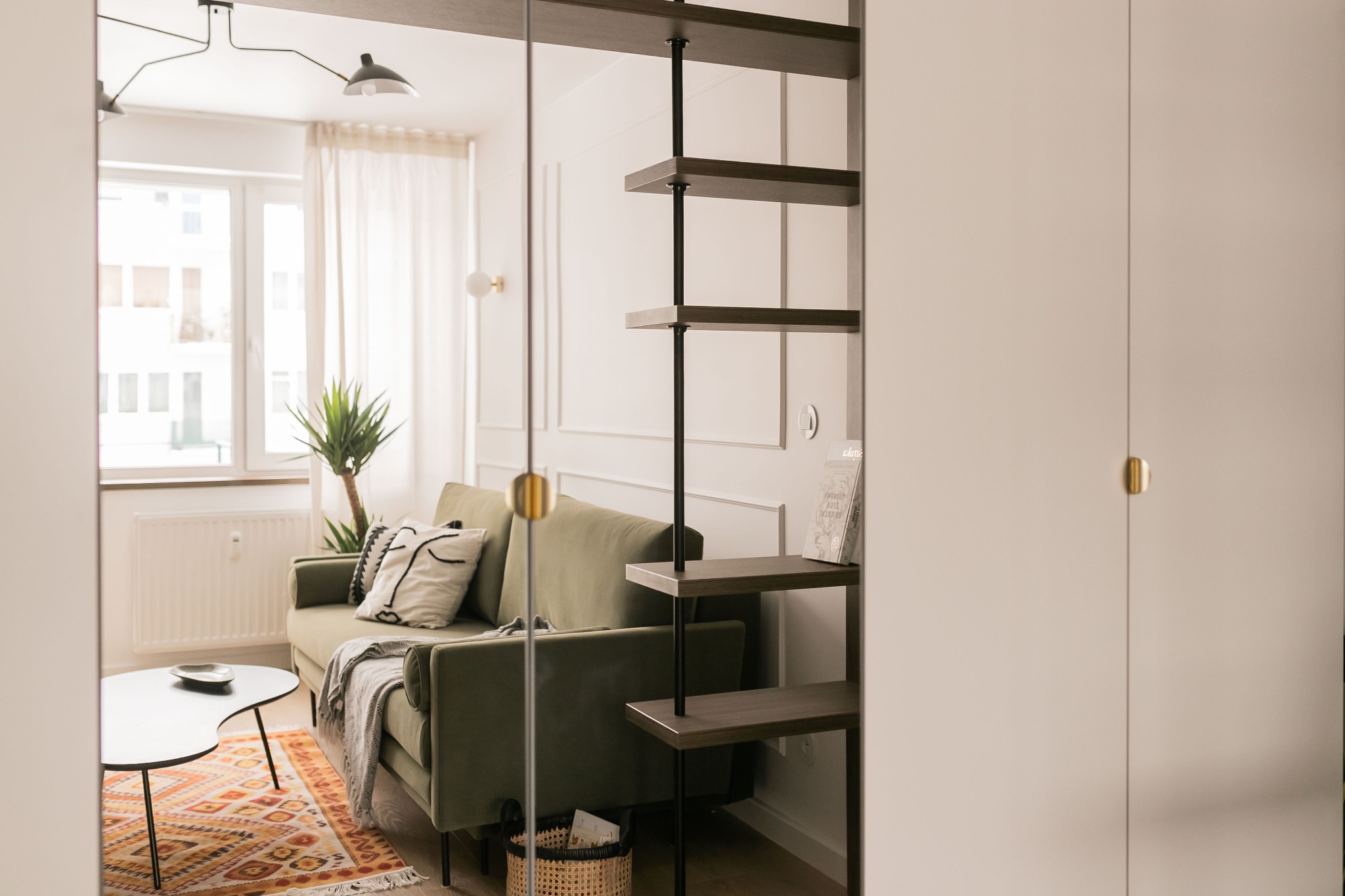 Salon w stylu nowoczesnym z zieloną sofą, wzorzystym dywanem i minimalistycznymi półkami, widok z perspektywy wejścia.